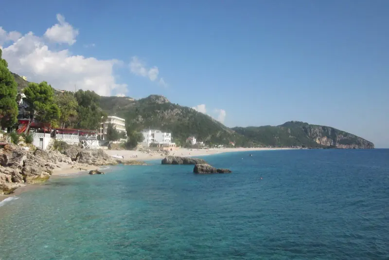 Clear green water and a pebble shore along the coast at Dhermi below forested mountains