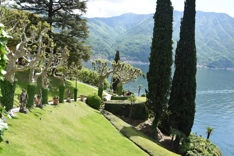 Lawns and clipped plane trees in the gardens of Villa del Balbianello above the lake with mountains beyond