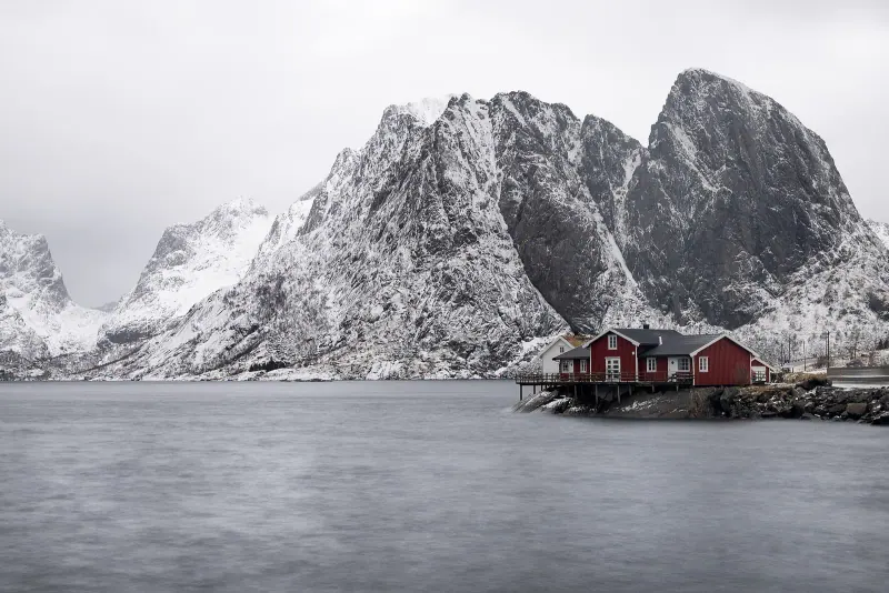 Lone red cabin on the shore of a still fjord below snow dusted Lofoten peaks