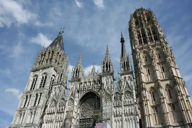 Rouen Cathedral with its spires and tower rising above the rooftops under a blue sky with clouds