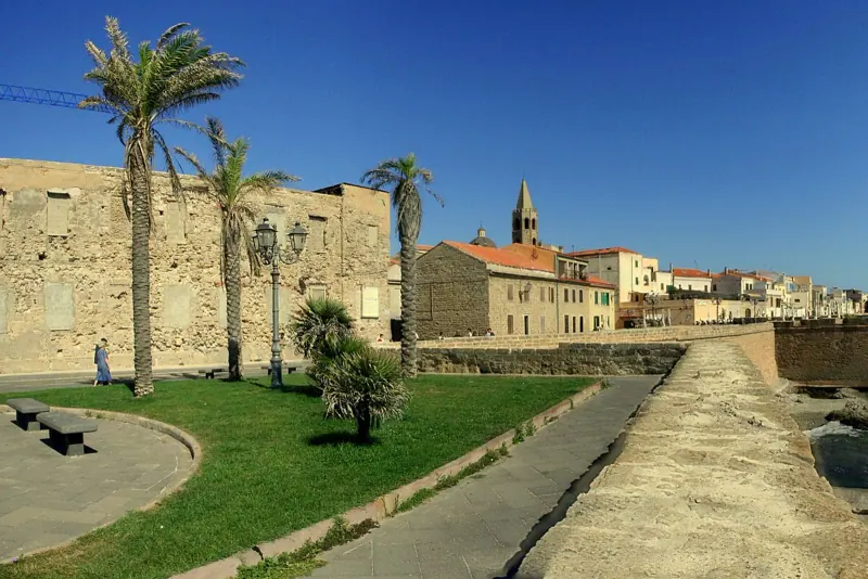 Alghero old town ramparts and church bell tower beside the sea, with palms and a green lawn