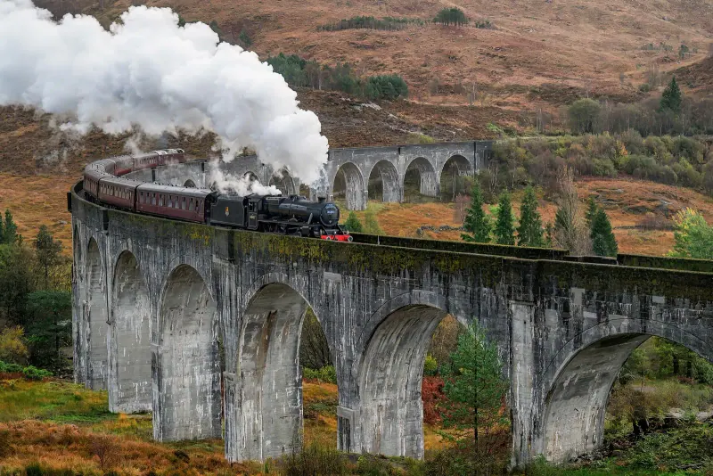 Steam train with a white plume curving across the arches of the Glenfinnan Viaduct